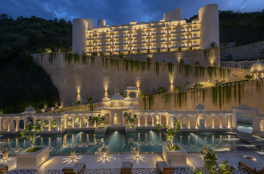 The resort sits like a fortress above Udaipur — floodlit at night, with a pool landscape and water features in the foreground.