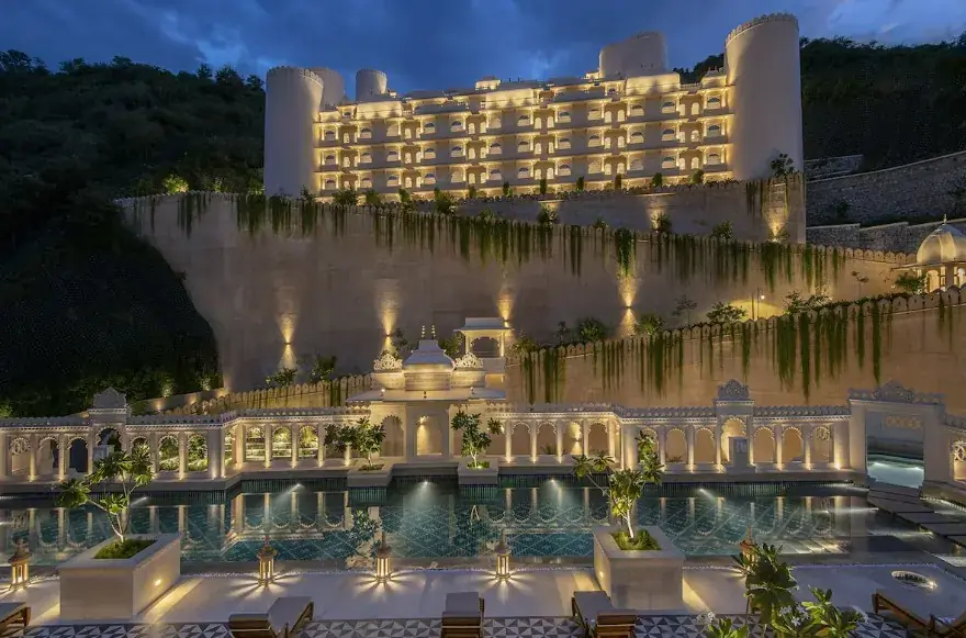 The resort sits like a fortress above Udaipur — floodlit at night, with a pool landscape and water features in the foreground.