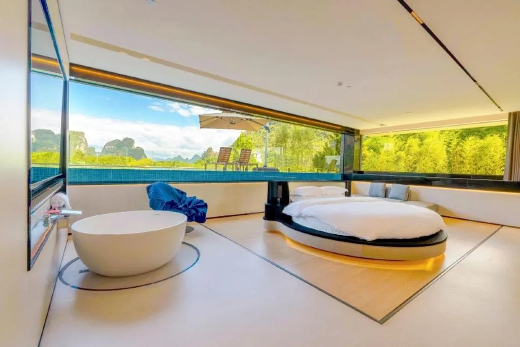 Rundes Bett und freistehende Wanne vor großflächiger Verglasung mit Blick auf Pool und Berge.