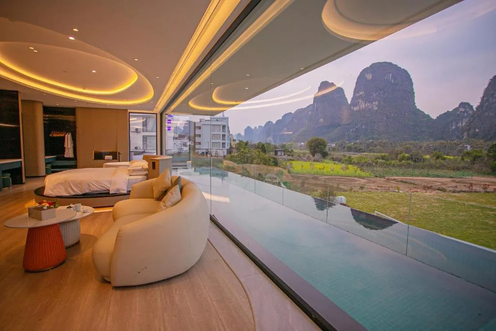 Rundes Bett und helles Sofa vor großer Glasfront mit Pool und Karstbergen in der Abenddämmerung.
