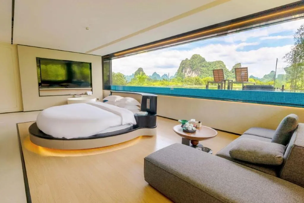 Rundes Bett und geschwungenes Sofa vor raumhoher Glasfront mit Pool und Karstbergen.