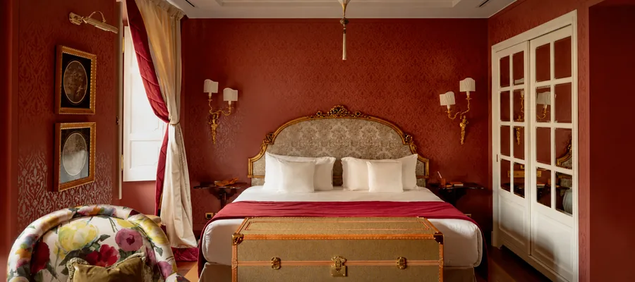 Schlafzimmer der Villa mit rot gemusterten Wänden, vergoldetem Bettkopf und antikem Reisekoffer